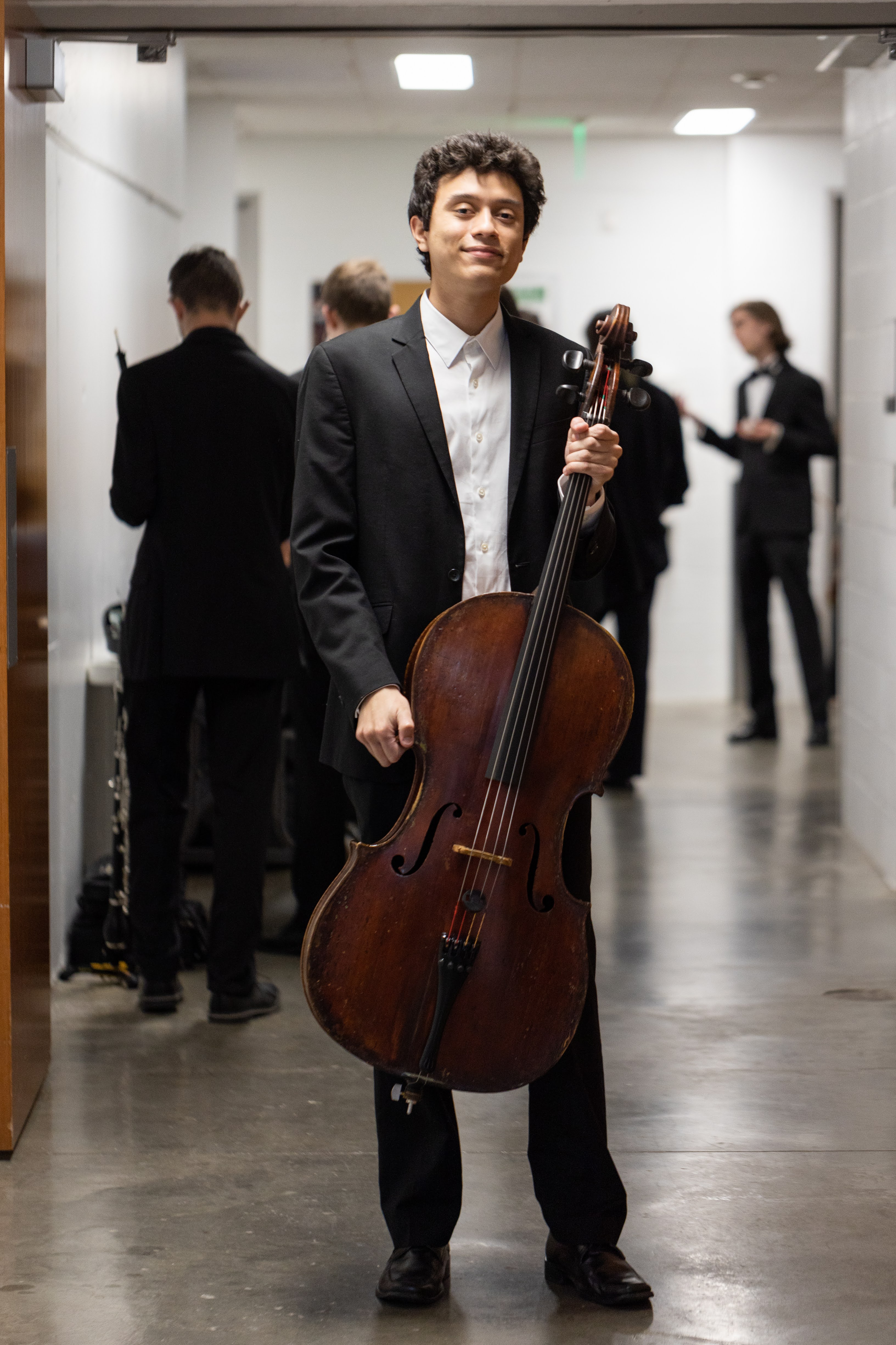 Andrés Pérez Rangel holding a cello in a hallway, with people in the background. ; Undergraduate Degree Recital: Andr&eacute;s P&eacute;rez Rangel, cello