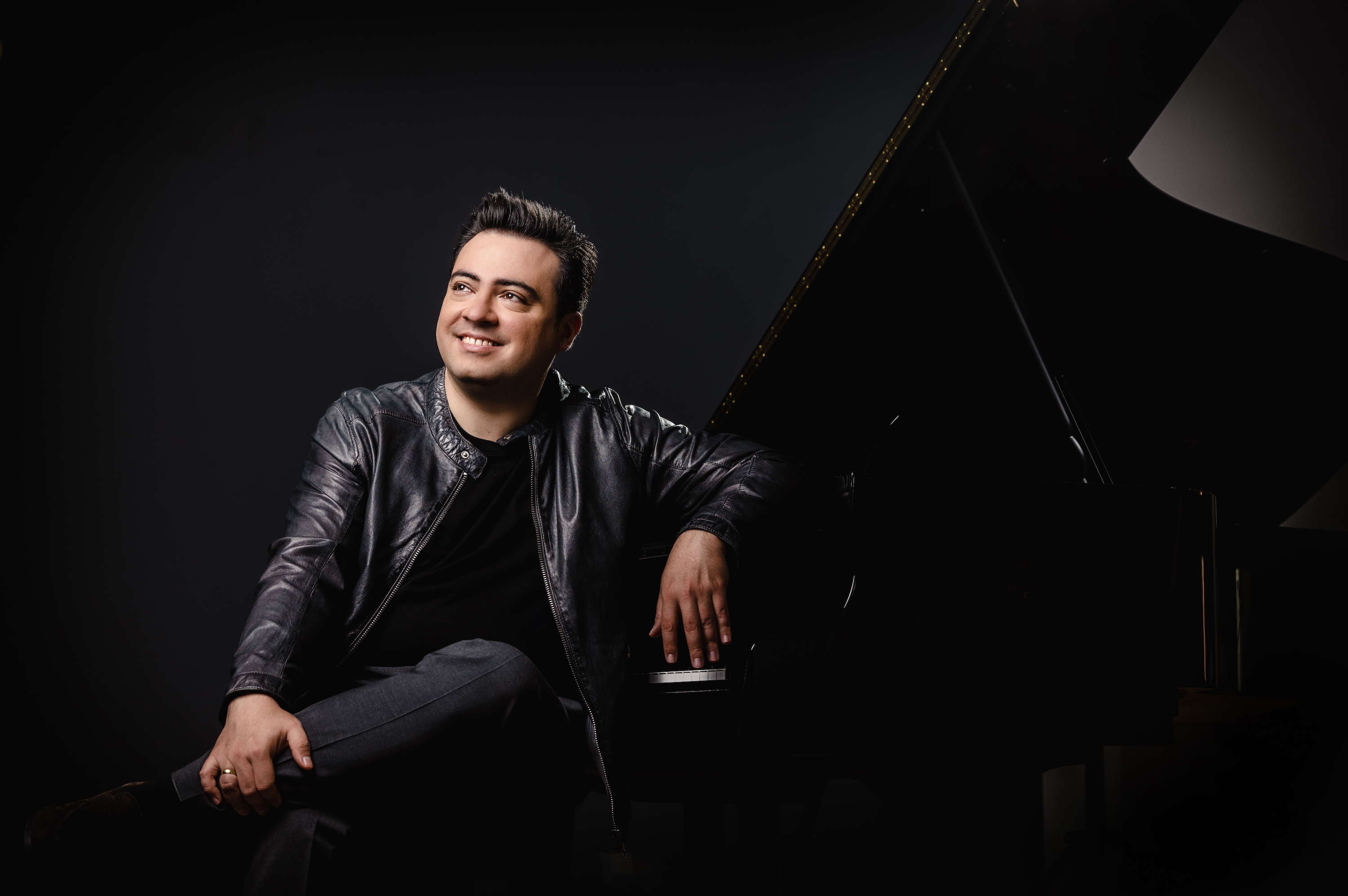 János Balázs leaning on a grand piano and smiling into the distance. ; Guest Artist Recital &amp; Masterclass: J&aacute;nos Bal&aacute;zs, piano