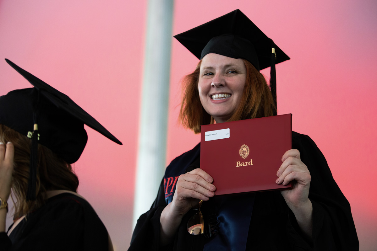 Smiling woman with red hair in cap and gown, holding up a red diploma for the camera.