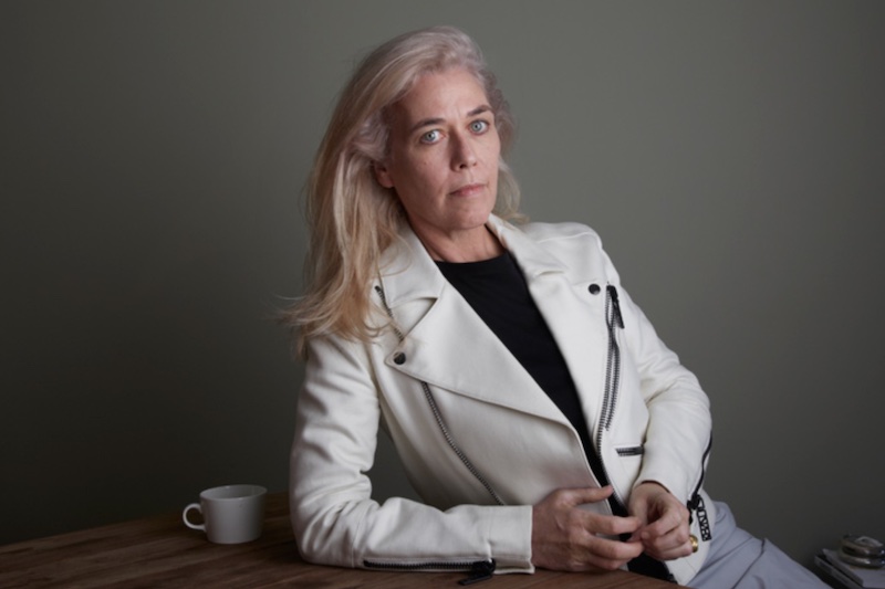 A woman with white, shoulder-length hair wearing a cream colored jacket with her hands folded in front of her looks directly into the camera. A mug sits to the left of her on the table on which she is leaning.