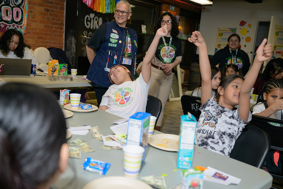 A group of young girls sitting at a table wearing Troop 6000 t-shirts raise their arms in the air and give the thumbs up.