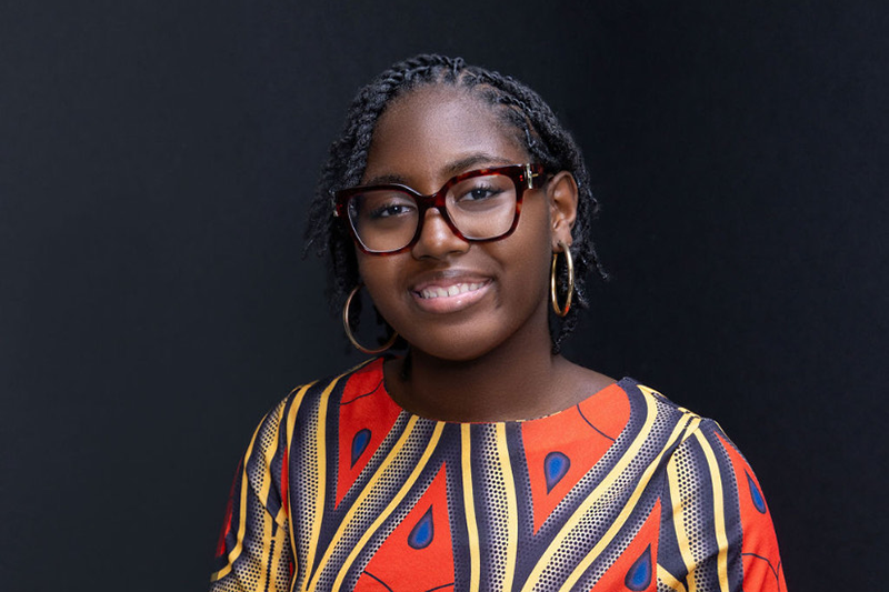 Rashanna James-Frison posing for a portrait in a colorful dress and black-framed glasses.