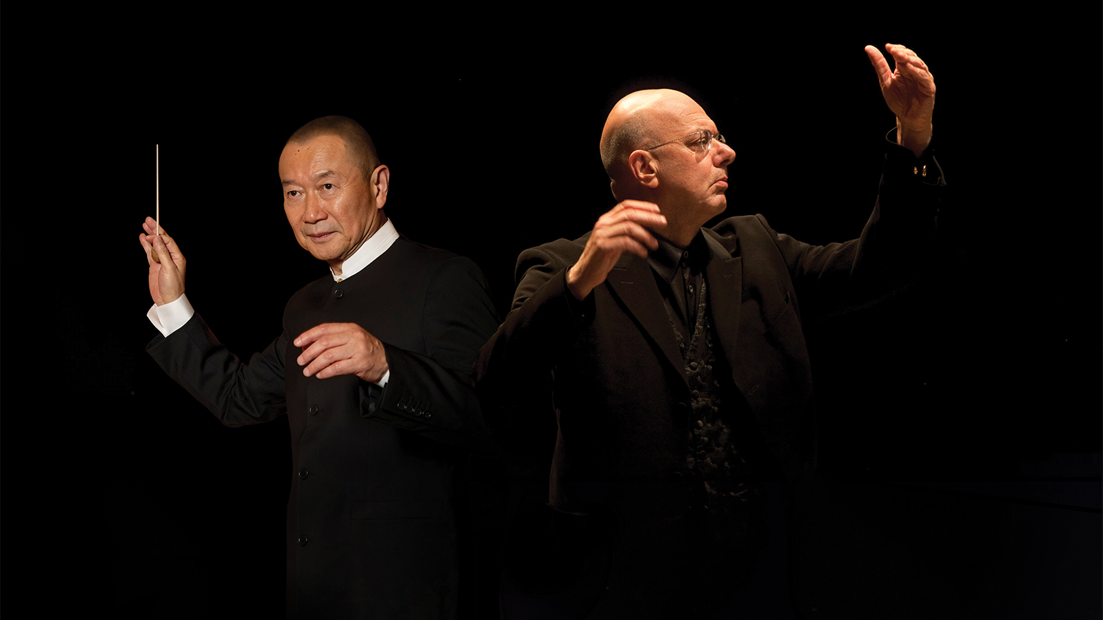 two men raise their hands to conduct against a black backdrop