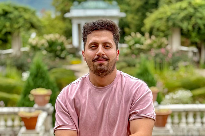 Man in pink t-shirt with moustache and light beard standing in front of formal gardens.