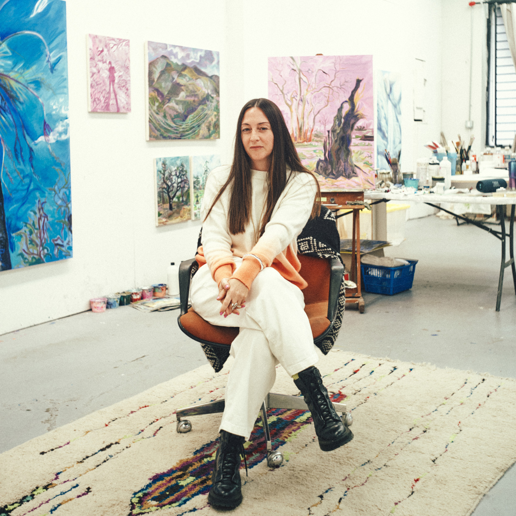 a woman in white with black boots sits in a studio surrounded by colorful paintings