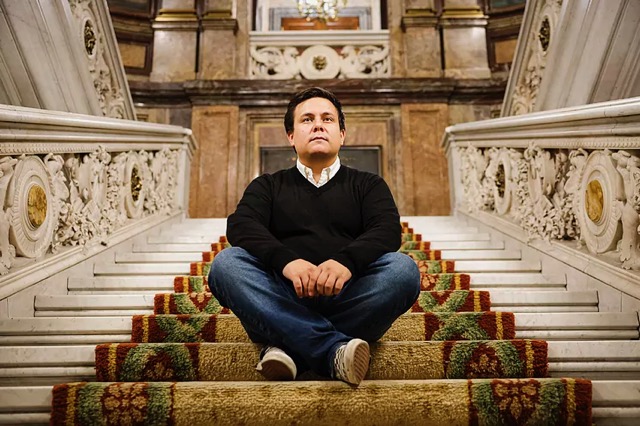 a man in jeans and a black sweater with button down white shirt sits on a tall set of stairs