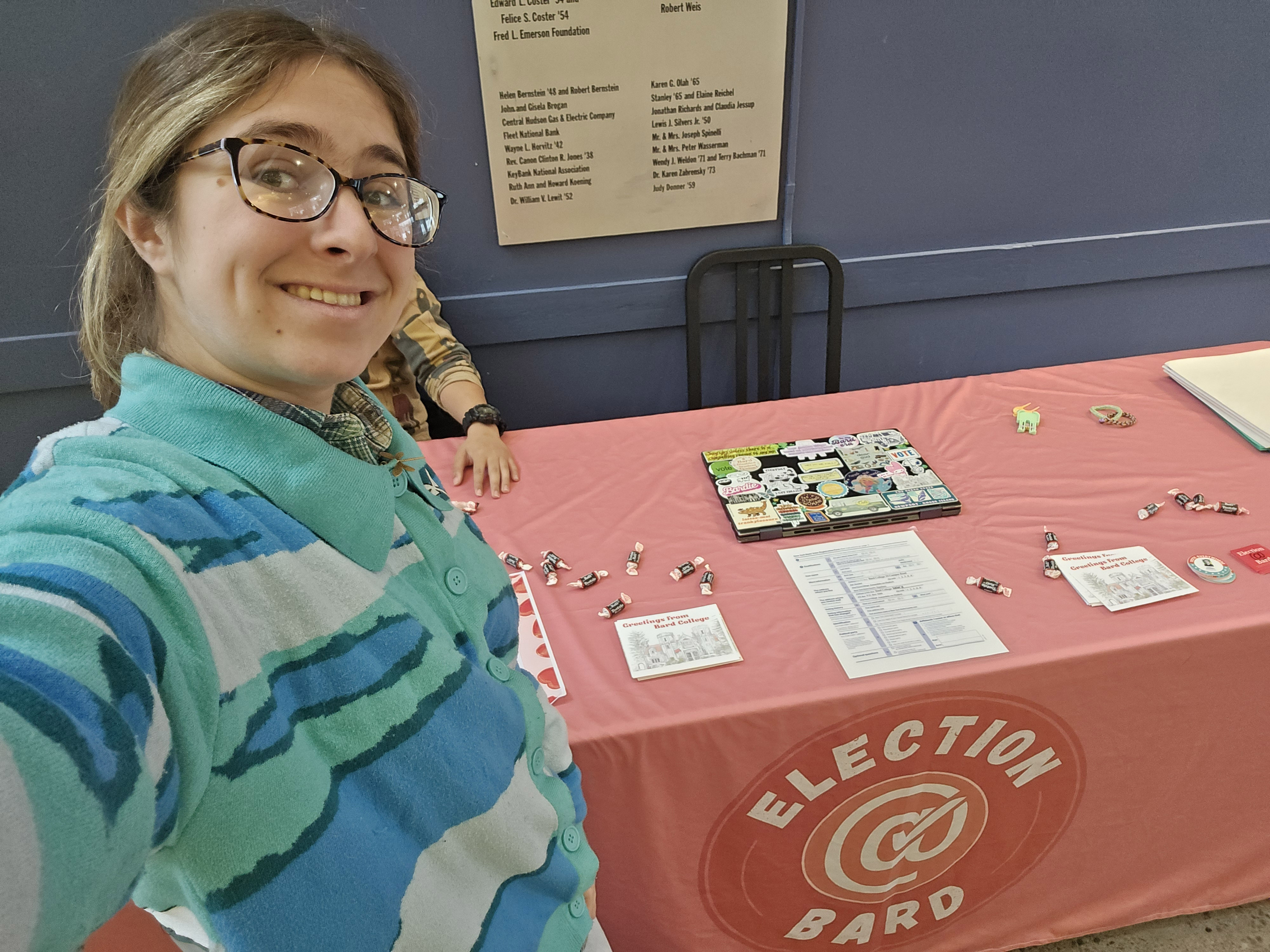 A student wearing a ponytail and striped shirt smiles in front of a voting booth