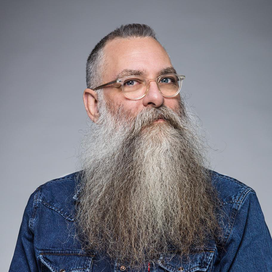 a bearded individual with glasses and a blue shirt looks at the viewer