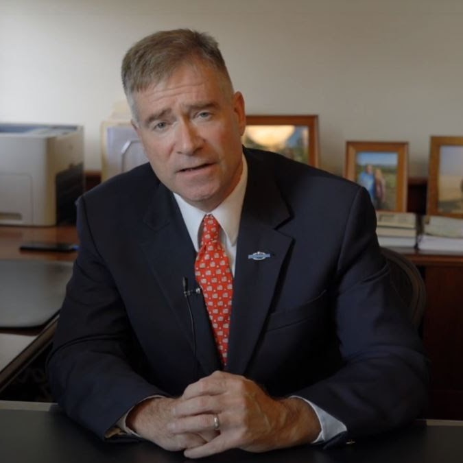 a man in a blue suit and red tie addresses the viewer from a desk