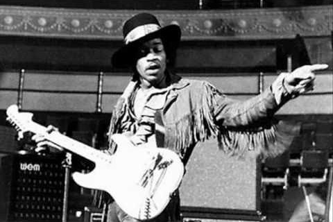A black and white photo of Jimi Hendrix in a fringed jacket, performing.