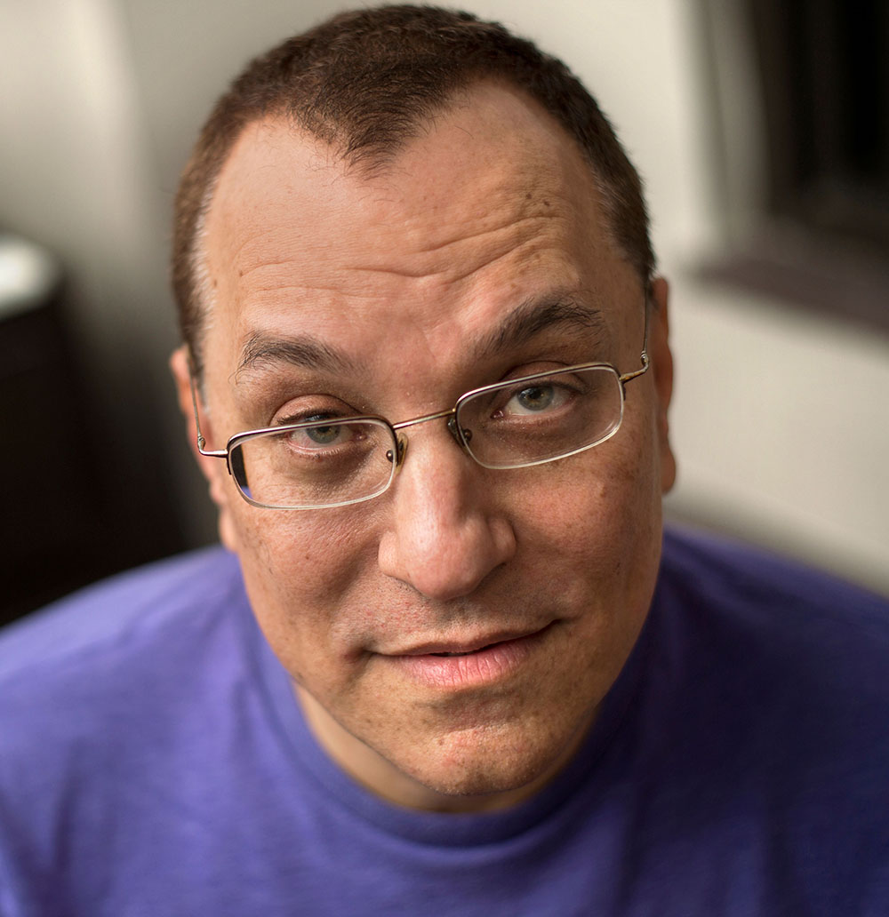 [Description: Close-up photograph of Kenny Fries, looking at the camera, wearing glasses
and a purple shirt.]