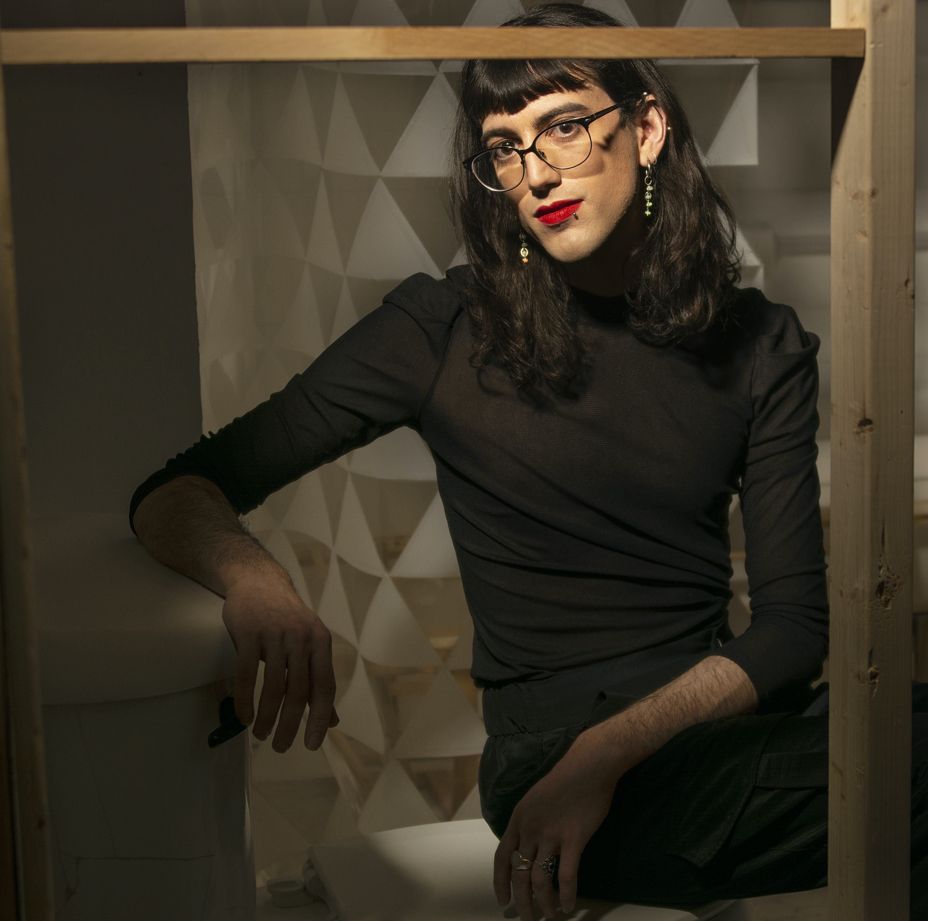 Figure seated with arm propped and looking at camera in black shirt with dark hair and bangs, wearing glasses and red lipstick.; Visit https://studioartvisitingartistopaleckerderuvo