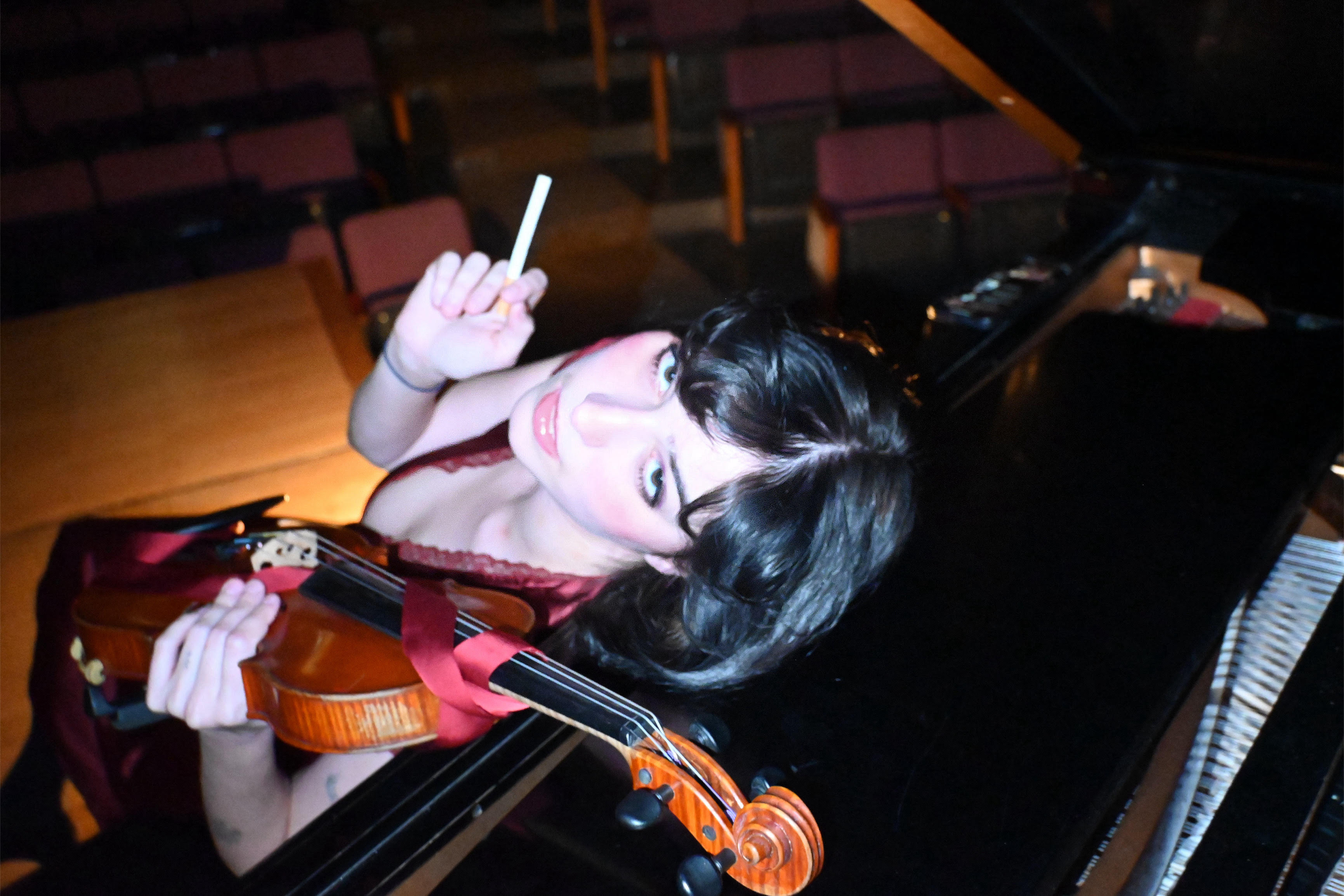 Klára Zaykova leaning over a piano, holding a violin and a cigarette. ; Undergraduate Degree Recital: Kl&aacute;ra Zaykova, violin