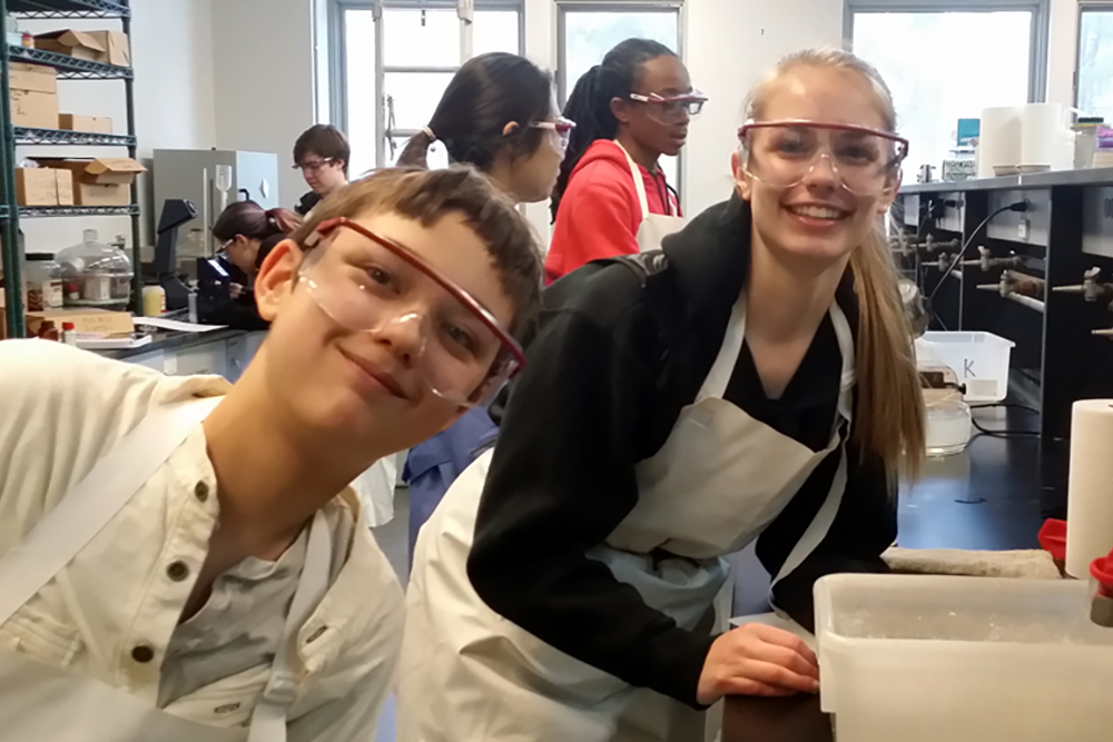 Two students in lab gear smile for the camera in a laboratory.